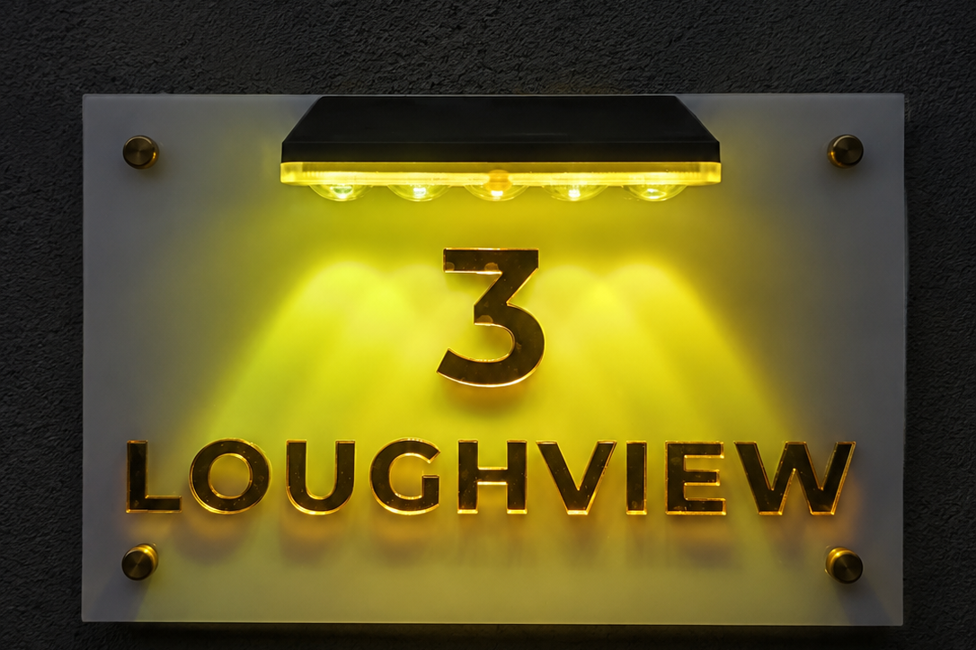 An illuminated house number sign reading '3 LOUGHVIEW' in gold lettering on a white rectangular panel, mounted against a dark textured wall with a built-in light fixture above.lumorastudio.ie lumora craft studio