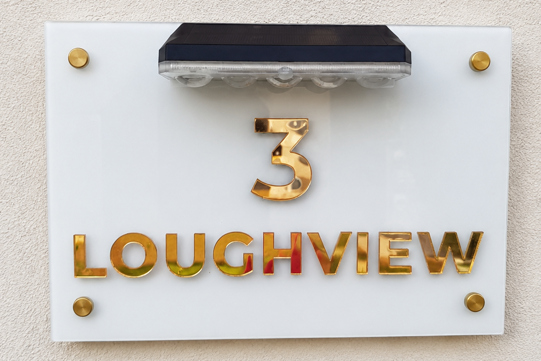 A white rectangular house number plaque mounted on a beige textured wall, featuring the gold metallic number 3 above the gold metallic text LOUGHVIEW, with a small black solar panel at the top.lumorastudio.ie lumora craft studio