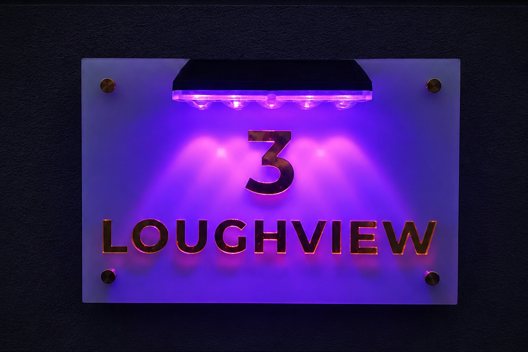A modern acrylic house number sign displaying '3 LOUGHVIEW' in gold lettering, illuminated by a purple LED light bar at the top against a dark wall.lumorastudio.ie lumora craft studio