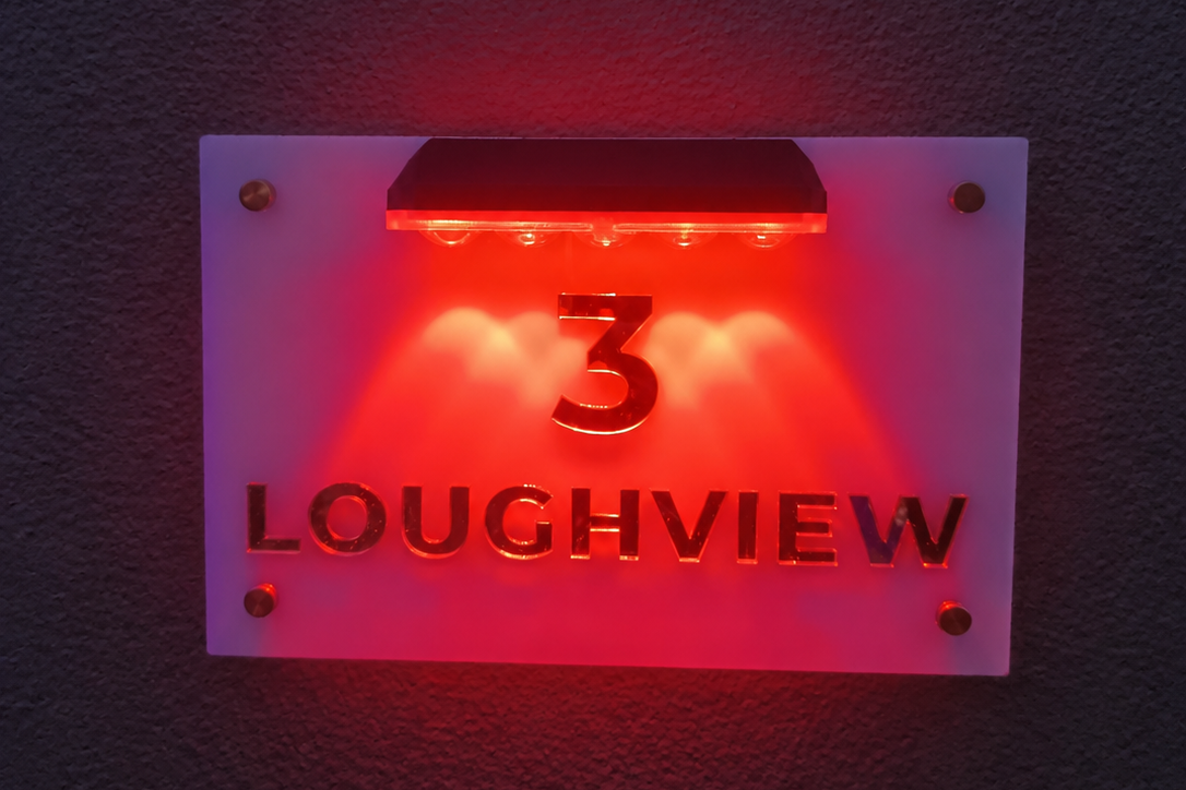 A modern house number light sign displaying '3 LOUGHVIEW' in red LED light on a frosted white panel, mounted on a dark textured wall.lumorastudio.ie lumora craft studio