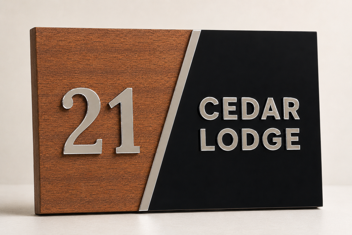 A rectangular house number sign with a diagonal split design: warm brown wood on the left showing the raised silver number 21, and black on the right with the silver text 'CEDAR LODGE'.lumorastudio.ie lumora craft studio