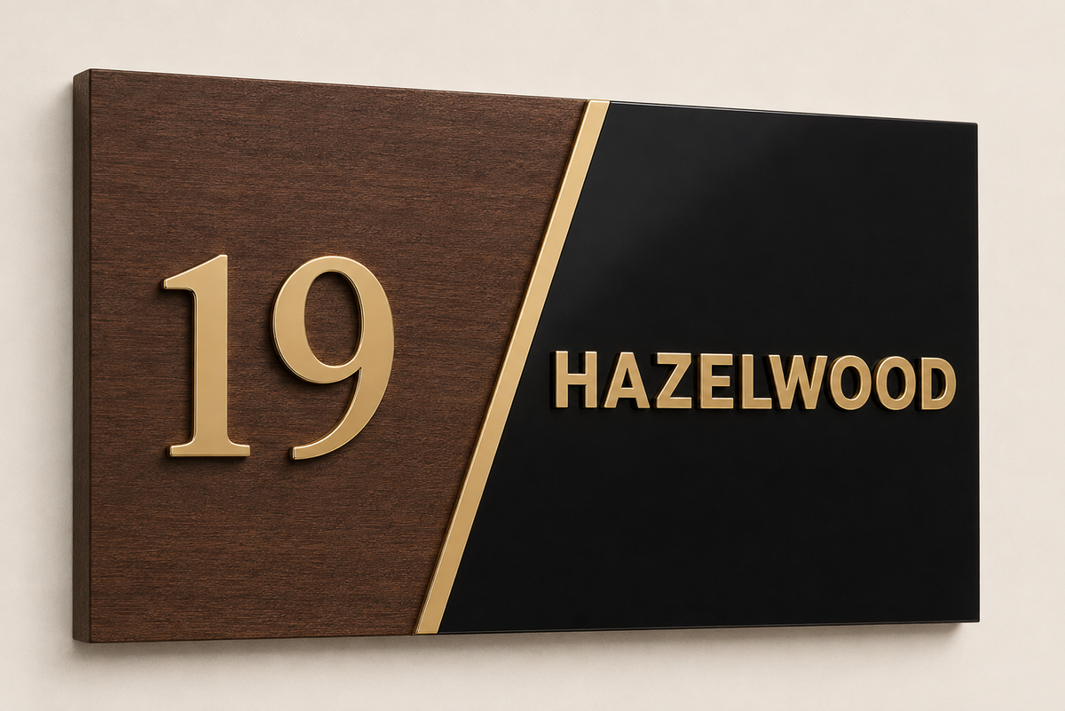 A modern house number and street name plaque mounted on a light wall. The left half is dark wood grain with raised gold numerals '19', and the right half is glossy black with raised gold text reading 'HAZELWOOD', divided by a diagonal gold stripe.lumorastudio.ie lumora craft studio