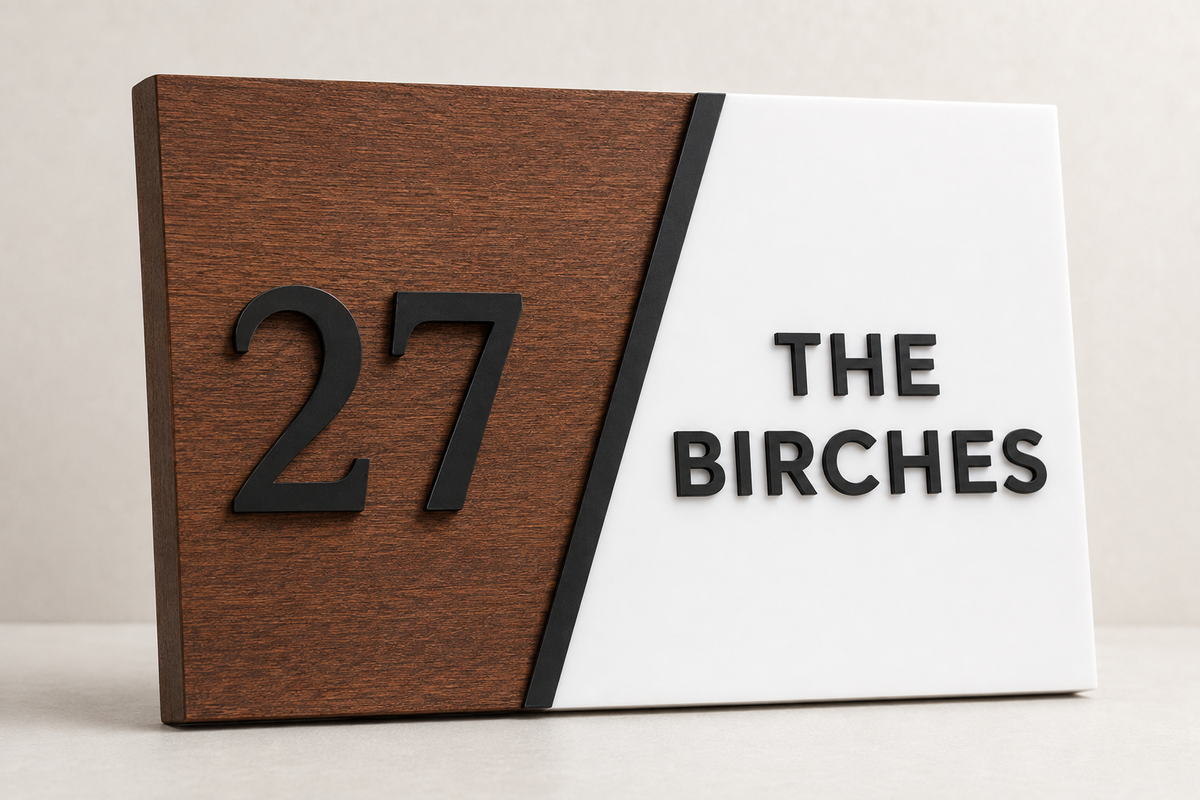 A rectangular house number sign with a split design: the left half is dark brown wood with raised black '27', and the right half is white with raised black 'THE BIRCHES'.lumorastudio.ie lumora craft studio