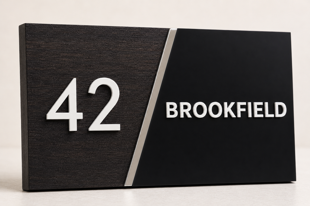 A modern house number sign displaying '42 BROOKFIELD' in white text on a split black and dark wood panel background with a thin metallic divider.lumorastudio.ie lumora craft studio