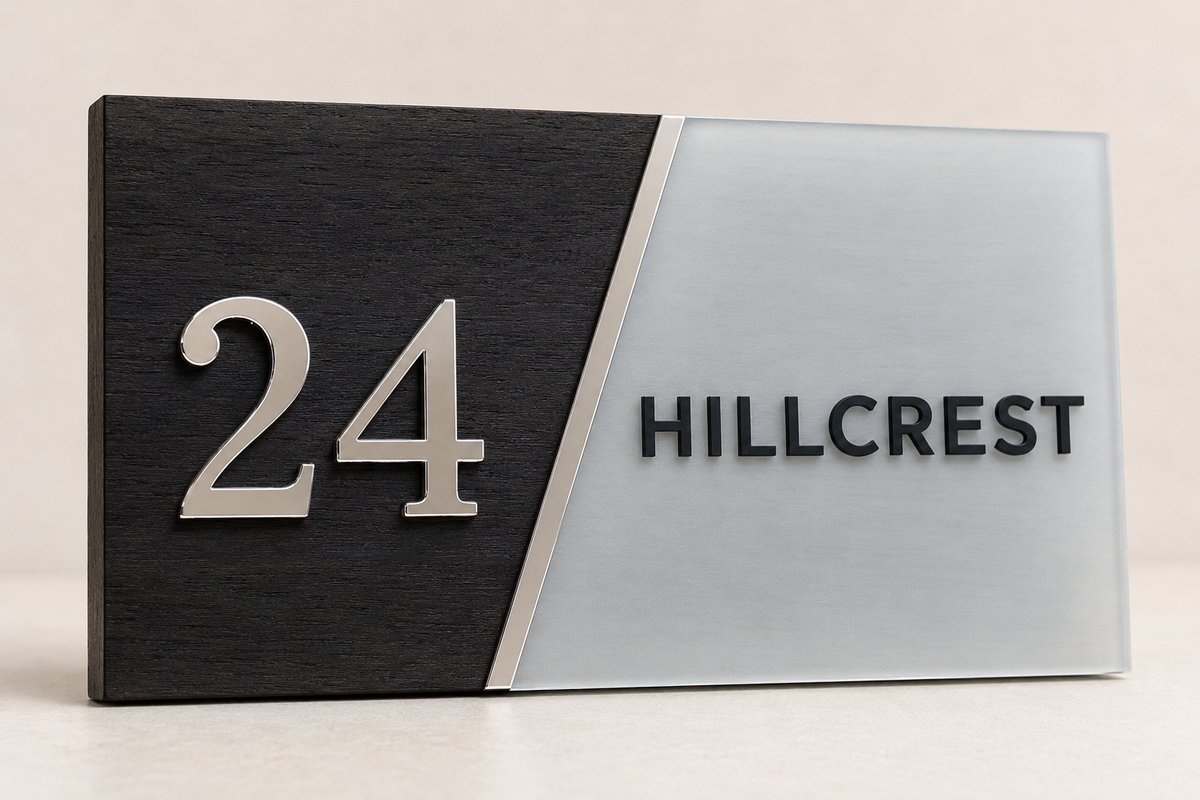 A modern house number and owner name plaque on a light beige background. The plaque is split diagonally into a dark wood-grain textured black section on the left with raised silver numbers '24', and a brushed silver section on the right with raised black letters spelling lumorastudio.ie lumora craft studio'HILLCREST'.