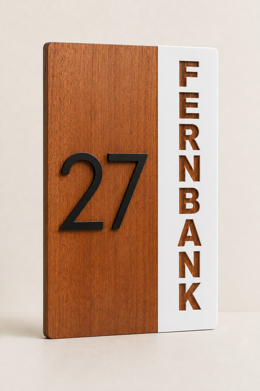 A modern house number plaque featuring a vertical split design with a warm brown wood grain panel on the left displaying the raised black number 27, and a white panel on the right with the word FERNBANK cut out in brown.Lumora Craft Studio, lumorastudio.ie