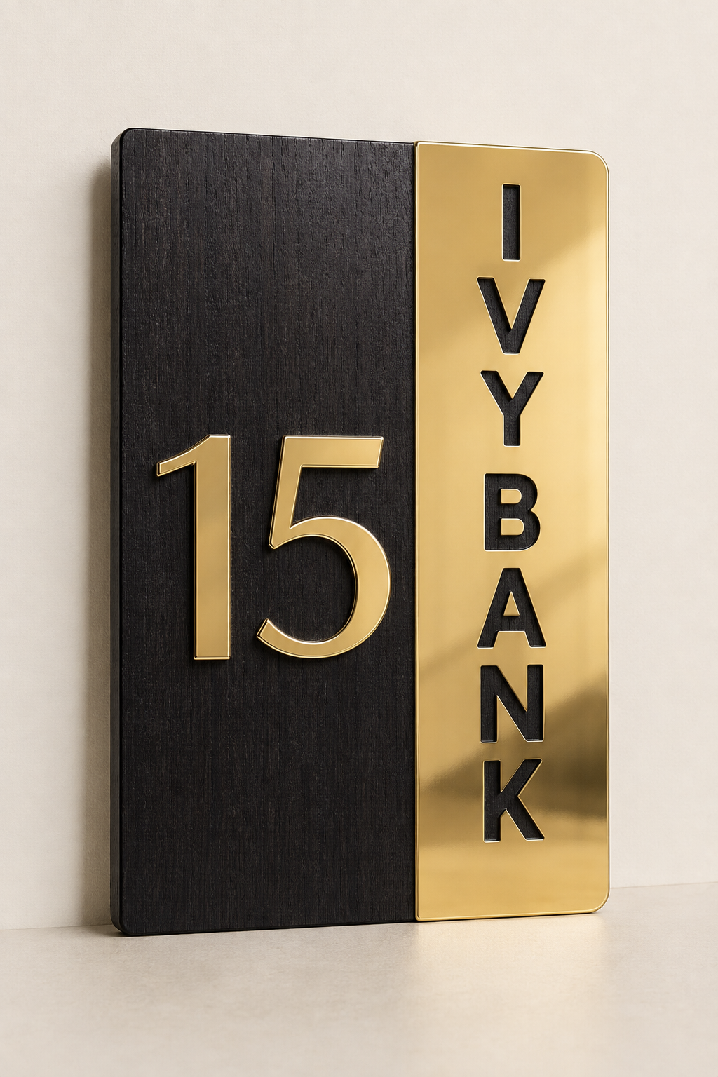 A modern house number sign featuring a split design with a dark wood-textured black left panel displaying the gold number 15, and a polished gold right panel with the words 'IVY BANK' in black vertical letters, shown against a neutral off-white background.