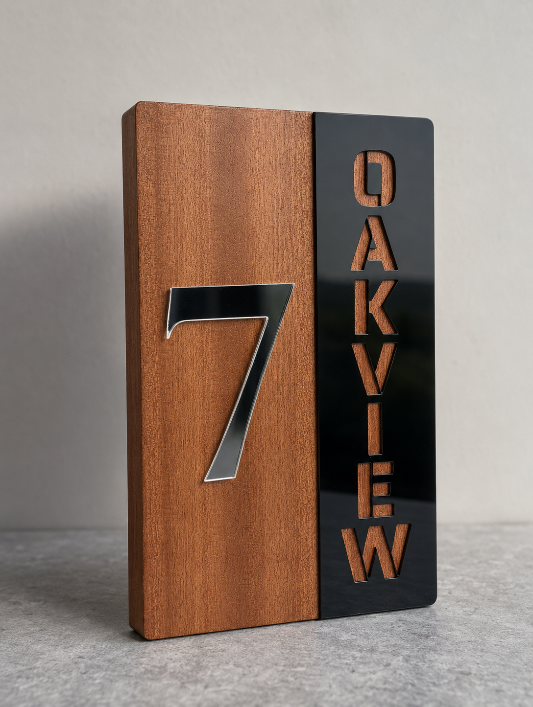 A modern rectangular house number sign with a split design: the left half is warm medium-brown wood with a large metallic silver number 7, and the right half is glossy black with the words 'OAKVIEW' in cutout letters showing the brown wood underneath. The sign stands on a gray surface against a light gray wall.Lumora Craft Studio, lumorastudio.ie
