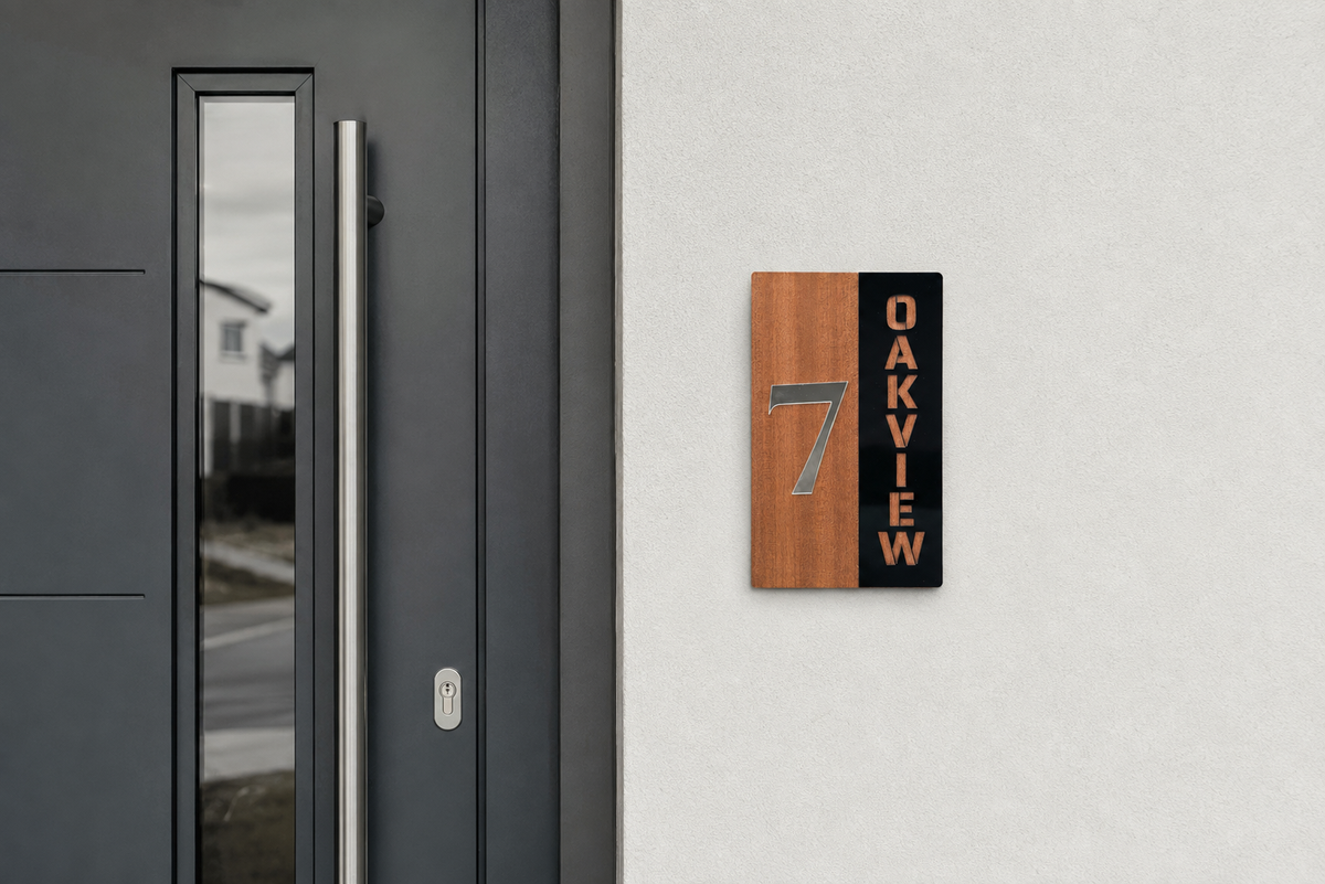 A modern house number sign mounted on a light gray exterior wall next to a dark gray front door. The sign features a vertical split design with a warm wood-grain left side displaying the number 7 in gray, and a black right side with the word OAKVIEW in gold lettering.Lumora Craft Studio, lumorastudio.ie