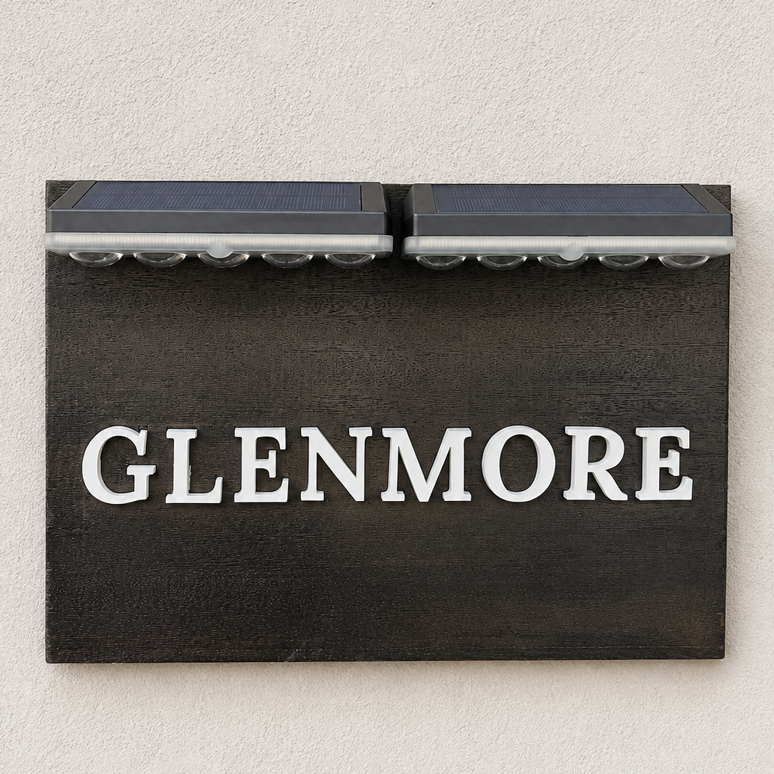 A rectangular black house sign with the name 'GLENMORE' in large white letters, mounted on a textured off-white wall. Two small dark solar panel strips are attached above the sign.lumorastudio.ie lumora craft studio