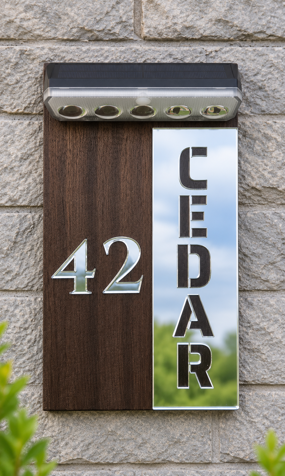 A modern house number and name sign mounted on a stone wall. The sign has a split design: the left half is dark brown wood with silver metallic '42' lettering, and the right half is a reflective silver mirror showing 'CEDAR' vertically with a sky and greenery reflection.Lumora Craft Studio, lumorastudio.ie