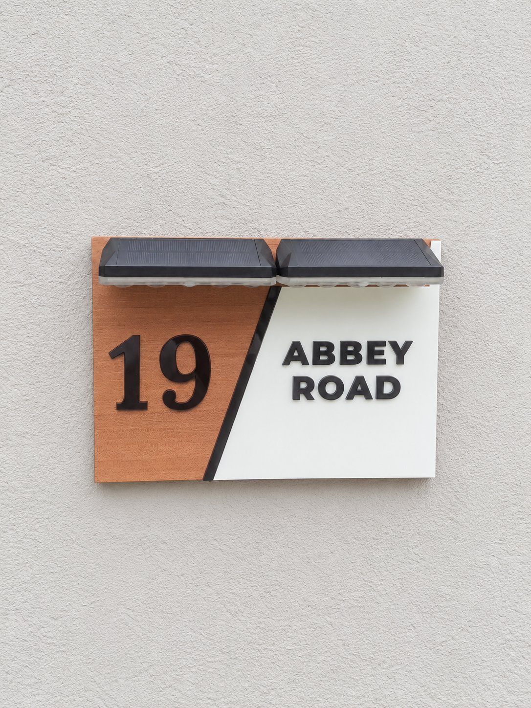 A modern split-color house number plaque mounted on a textured light gray wall. The left half is a warm wood tone with the black number '19', and the right half is white with the black text 'ABBAY ROAD'.lumorastudio.ie lumora craft studio