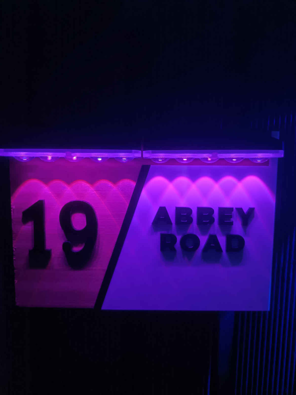 A rectangular illuminated sign with the number 19 on the left and the words ABBEY ROAD on the right, lit in pink and purple tones against a dark background.Personalised diagonal wood and acrylic house plaque with raised lettering