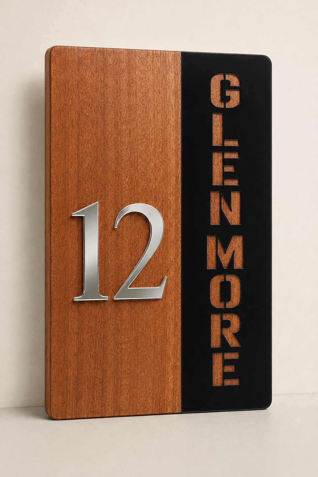 A rectangular wooden house number plaque with a split design: warm medium-brown wood on the left side featuring a silver metallic '12', and black on the right side with vertical gold-toned 'GLENMORE' lettering. The plaque is photographed standing upright against a plain light beige wall and surface.