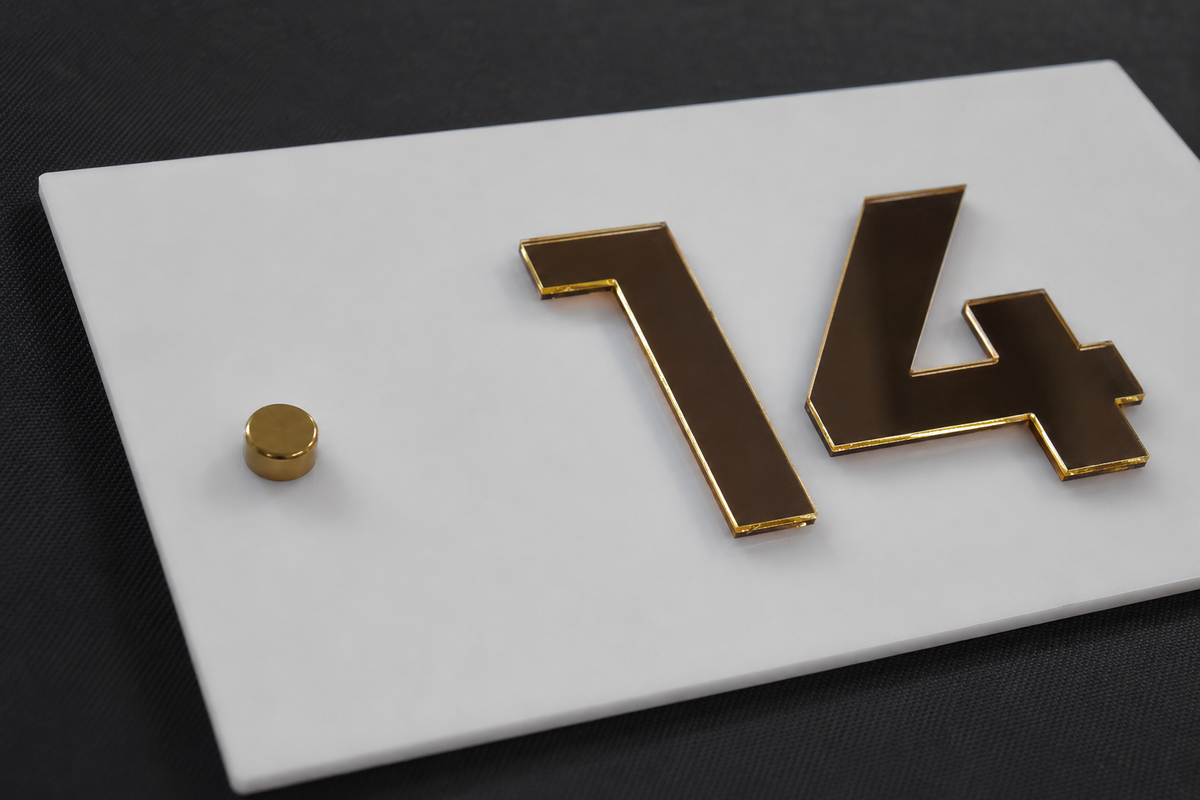 A close-up of a white rectangular plaque or sign with raised gold numerals '14' and a small gold circular accent, photographed on a dark textured background.lumorastudio.ie lumora craft studio