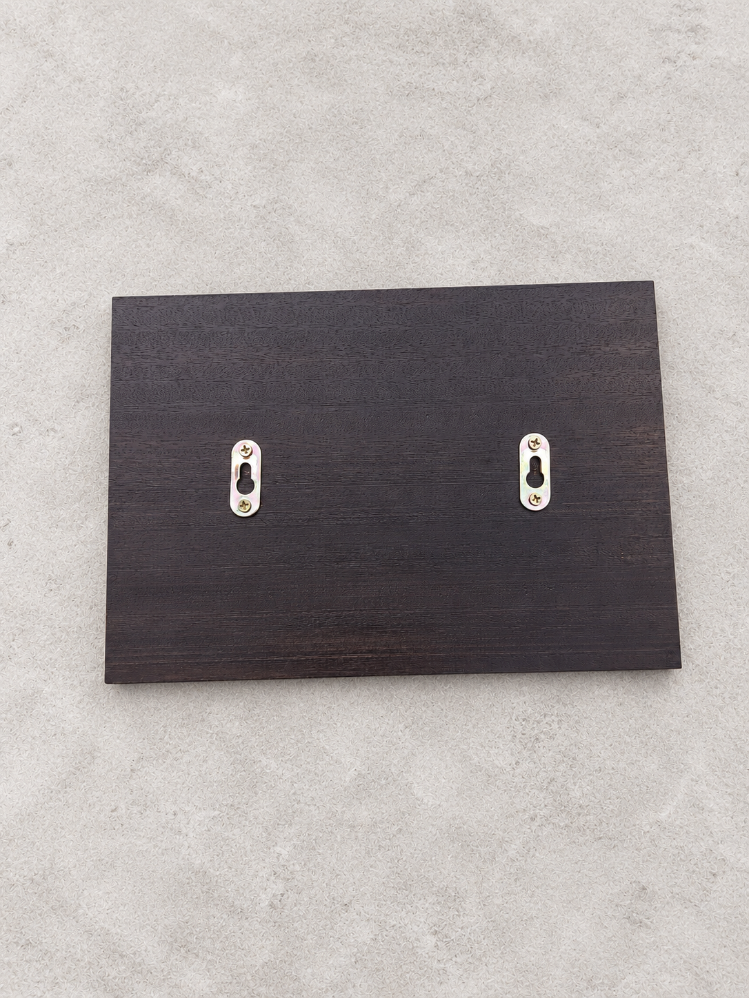 A dark brown wooden rectangular board with two silver metal keyhole-shaped wall hooks, shown against a textured light gray background.lumorastudio.ie lumora craft studio