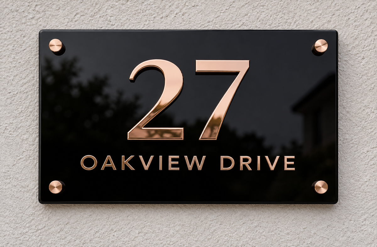 A rectangular black house number plaque mounted on a textured light gray wall. The plaque features the number 27 in large metallic rose-gold/copper letters above the words OAKVIEW DRIVE in matching metallic lettering, secured with four corner screws.lumorastudio.ie lumora craft studio