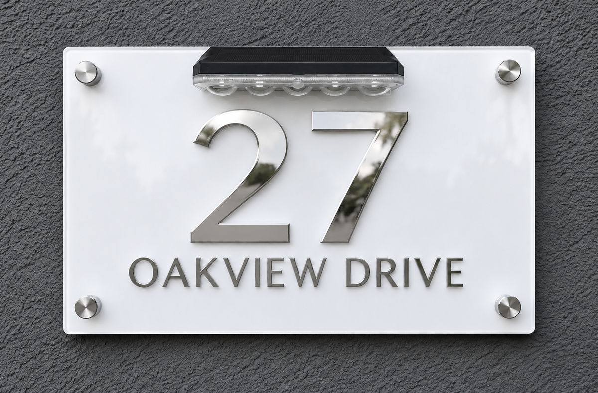 A modern house number sign reading '27 OAKVIEW DRIVE' on a white rectangular plaque with silver metal numerals and lettering, mounted against a dark textured wall. A small solar panel is attached above the numbers.lumorastudio.ie lumora craft studio
