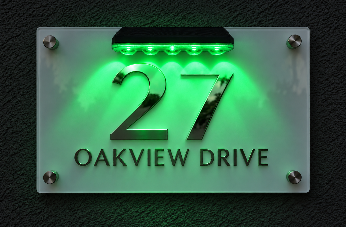 A modern rectangular glass house number sign mounted on a dark textured wall. The sign features the number 27 in polished chrome, with the text 'OAKVIEW DRIVE' below in dark lumorastudio.ie lumora craft studiolettering.