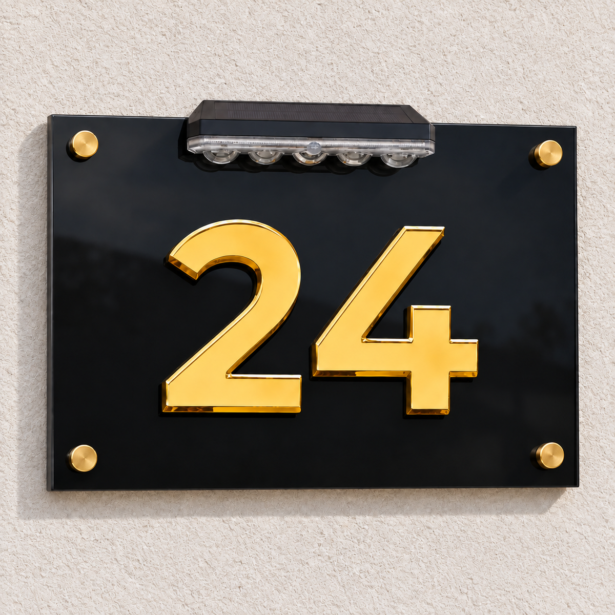 A modern house number plaque featuring the number 24 in polished gold on a glossy black background, mounted on a textured beige wall with a built-in solar light at the top.lumorastudio.ie lumora craft studio