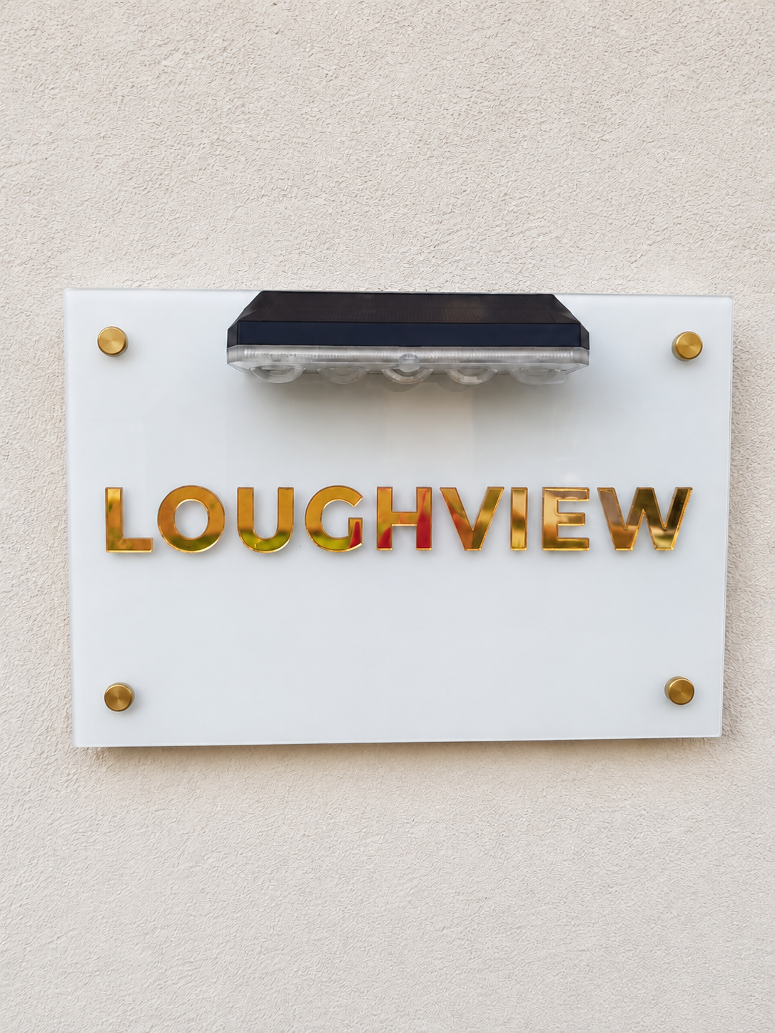 A white rectangular wall sign with the word 'LOUGHVIEW' in raised gold letters, mounted on a beige textured wall. A small black solar light fixture is mounted above the sign.lumorastudio.ie lumora craft studio